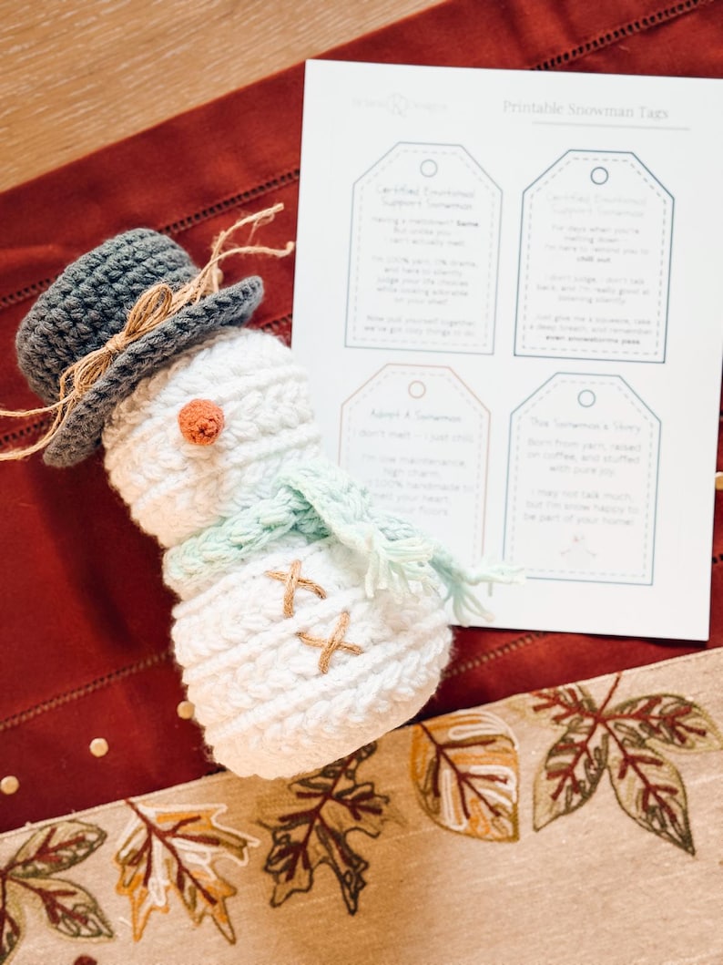 White crochet snowman with gray hat lying beside a sheet of printable snowman tags on a red table runner