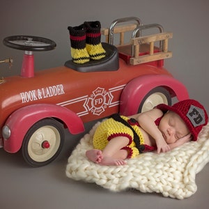 Könnte beinhalten: Ein neugeborenes Baby, das ein rot-gelbes Feuerwehrkostüm trägt, schläft auf einer weißen Strickdecke neben einem roten Spielzeugfeuerwehrauto. Das Auto hat eine Hakenleiter und die Worte "Hook & Ladder" sind an der Seite aufgedruckt. Das Baby trägt eine rote Feuerwehrkappe mit den Buchstaben "FD" darauf.