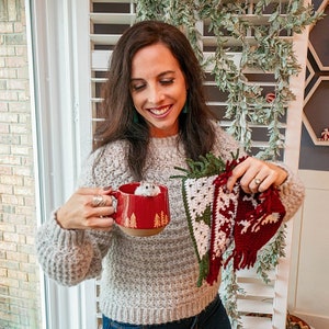 May include: A woman wearing a gray sweater holds a red mug with a small white hamster inside. She is also holding three crocheted Christmas ornaments, one green, one white and green, and one red.