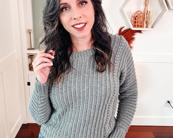 May include: A woman wearing a gray knitted sweater with a ribbed texture and blue jeans. She is standing in front of a white wall with a wooden shelf behind her.
