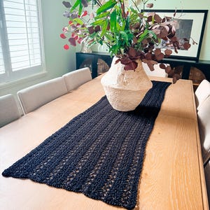 A cream-colored vase with red and green foliage sits on a wooden table with a dark woven table runner, creating the perfect setting for displaying unique jewellery or presenting a thoughtful handmade gift.