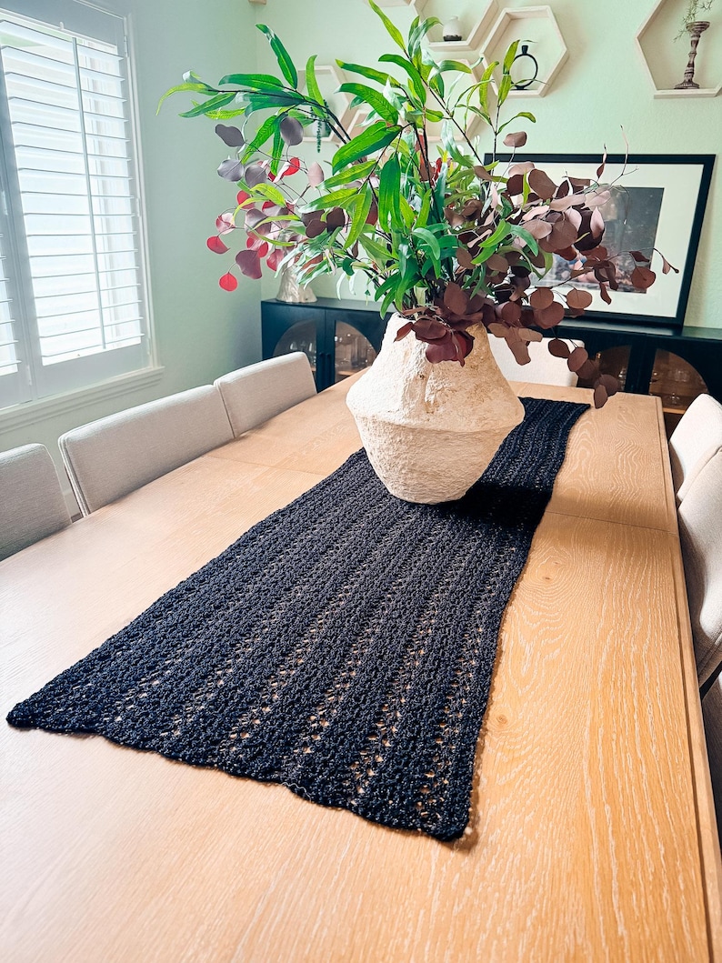 A cream-colored vase with red and green foliage sits on a wooden table with a dark woven table runner, creating the perfect setting for displaying unique jewellery or presenting a thoughtful handmade gift.