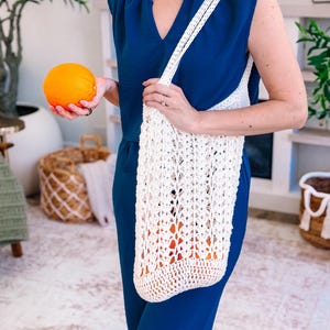 Puede incluir: Un bolso tote de crochet blanco de tejido abierto se lleva al hombro. La bolsa est&aacute; llena de naranjas. Una persona sostiene una naranja en la otra mano. La persona lleva un mono azul.