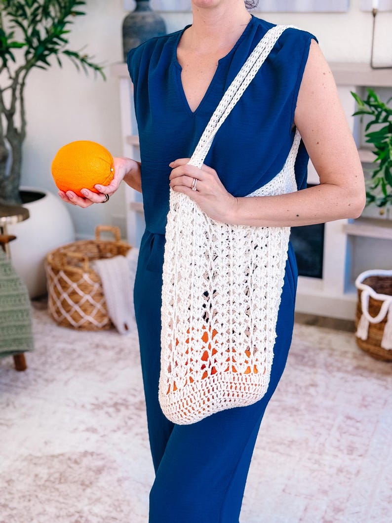 Puede incluir: Un bolso tote de crochet blanco de tejido abierto se lleva al hombro. La bolsa est&aacute; llena de naranjas. Una persona sostiene una naranja en la otra mano. La persona lleva un mono azul.