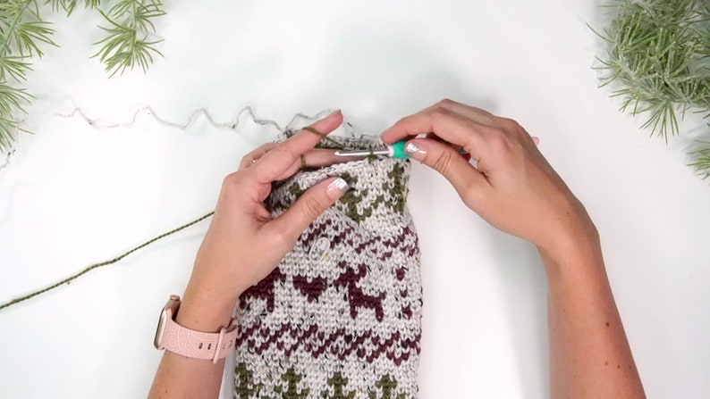 Op de afbeelding: Een haakproject in uitvoering met een fair isle patroon van rendier- en hartmotieven in olijfgroen en bordeauxrood op een gespikkelde haverkleurige achtergrond, gemaakt met een groenblauwe haaknaald en olijfgroen garen.