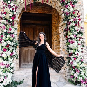 May include: A black floor-length dress with a sweetheart neckline and a high front slit, paired with a black crocheted shawl featuring an openwork pattern. The garment is worn in front of a stone archway decorated with white, pink, and magenta flowers.