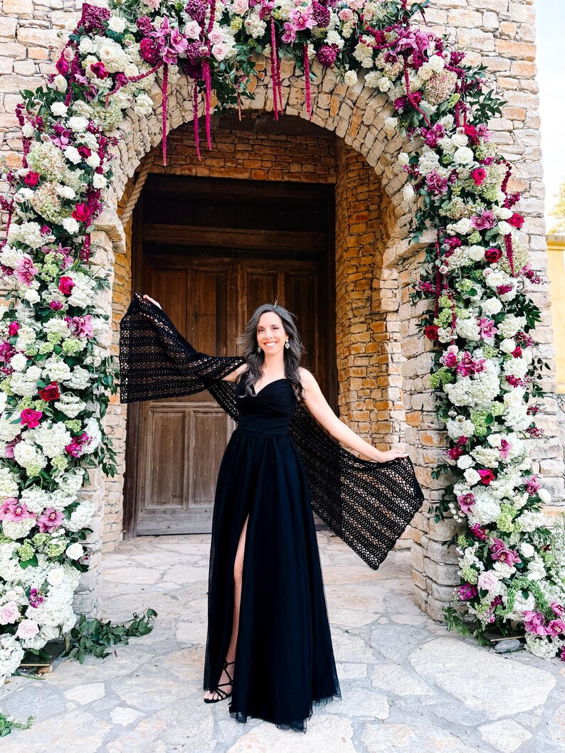 May include: A black floor-length dress with a sweetheart neckline and a high front slit, paired with a black crocheted shawl featuring an openwork pattern. The garment is worn in front of a stone archway decorated with white, pink, and magenta flowers.