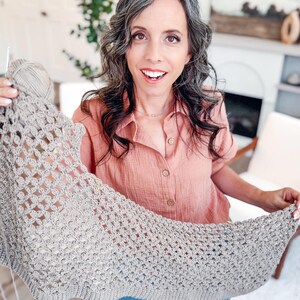 May include: A light beige crochet shawl featuring an intricate openwork pattern with a solid ribbed border. The textile is crafted from a neutral-toned yarn and is held up to display the detailed stitch work and the overall triangular shape.