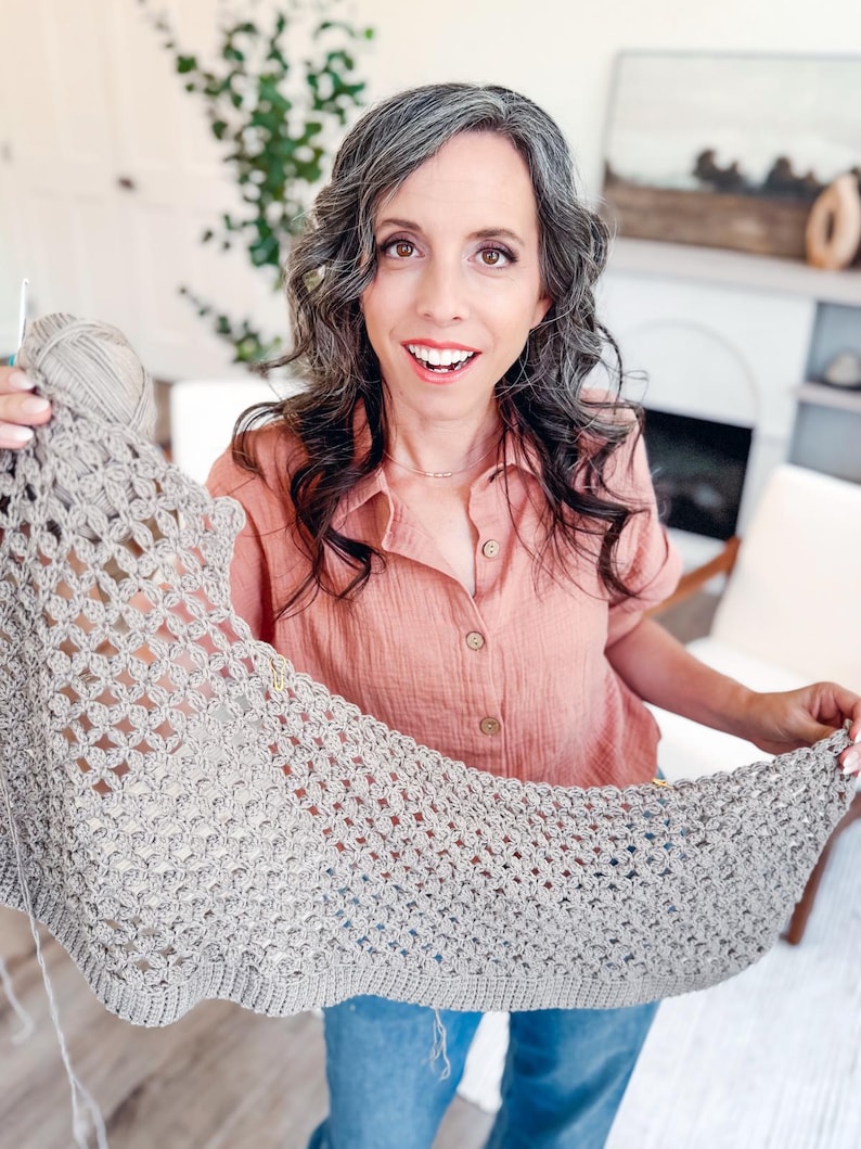 May include: A light beige crochet shawl featuring an intricate openwork pattern with a solid ribbed border. The textile is crafted from a neutral-toned yarn and is held up to display the detailed stitch work and the overall triangular shape.