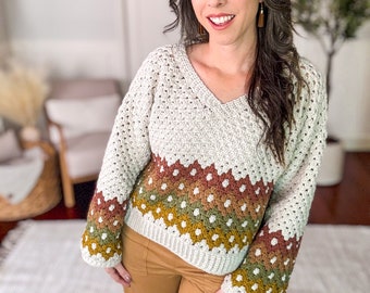 May include: A long-sleeve crochet sweater featuring a V-neckline and a ribbed hem. The upper portion is cream-colored, while the lower section and cuffs display a repeating geometric pattern in horizontal bands of rust, olive green, and mustard yellow.