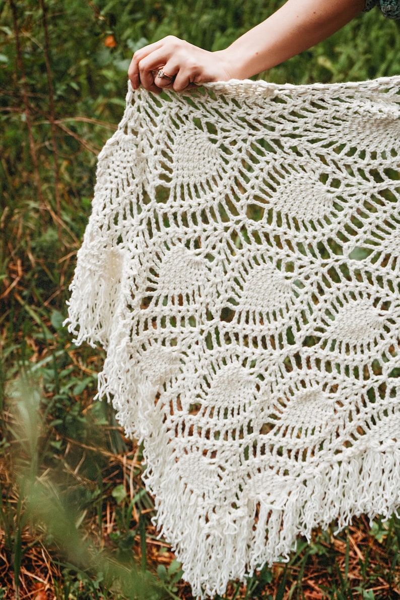 A woman wearing a white shawl decorated with the intricate crochet pineapple stitch, with fringed edges. She stands in an outdoor green space, surrounded by greenery and sunshine.