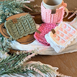 May include: A collection of handmade crochet mug cozies and coasters in various colors, including pink, green, and white. The cozies feature textured patterns and are adorned with small leather tags. A beige mug sits in a pink cozy. The image includes floral accents.