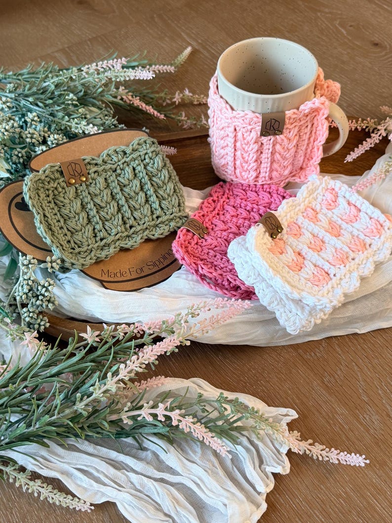May include: A collection of handmade crochet mug cozies and coasters in various colors, including pink, green, and white. The cozies feature textured patterns and are adorned with small leather tags. A beige mug sits in a pink cozy. The image includes floral accents.