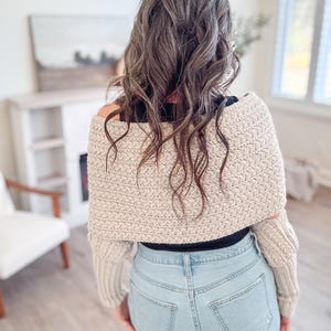A person with wavy brown hair in a beige crochet sweater, adorned with unique jewellery, stands in blue jeans in a room featuring a chair and an intriguing painting.