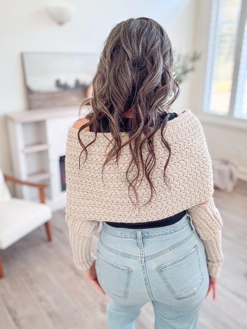 A person with wavy brown hair in a beige crochet sweater, adorned with unique jewellery, stands in blue jeans in a room featuring a chair and an intriguing painting.