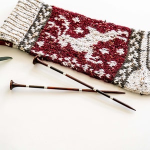 Puede incluir: Una media de Navidad de punto roja y blanca con un diseño de reno, junto con dos agujas de tejer de madera.