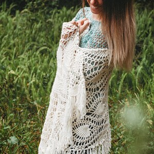 A woman wearing a white shawl decorated with the intricate crochet pineapple stitch, with fringed edges. She stands in an outdoor green space, surrounded by greenery and sunshine.