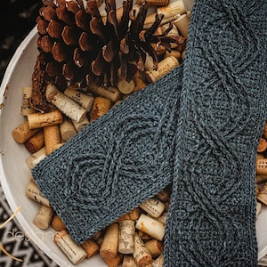 May include: A pair of blue knitted arm warmers with a cable knit pattern, displayed in a white bowl filled with wine corks and a pine cone.