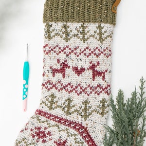 Op de afbeelding: Een gebreide kerstkous met een cr&egrave;mekleurige basis van gespikkeld garen, versierd met horizontale banden van donkerrode rendieren en olijfgroene dennenbomen, afgewerkt met een geribbelde olijfgroene boord en een bruine leren ophanglus.