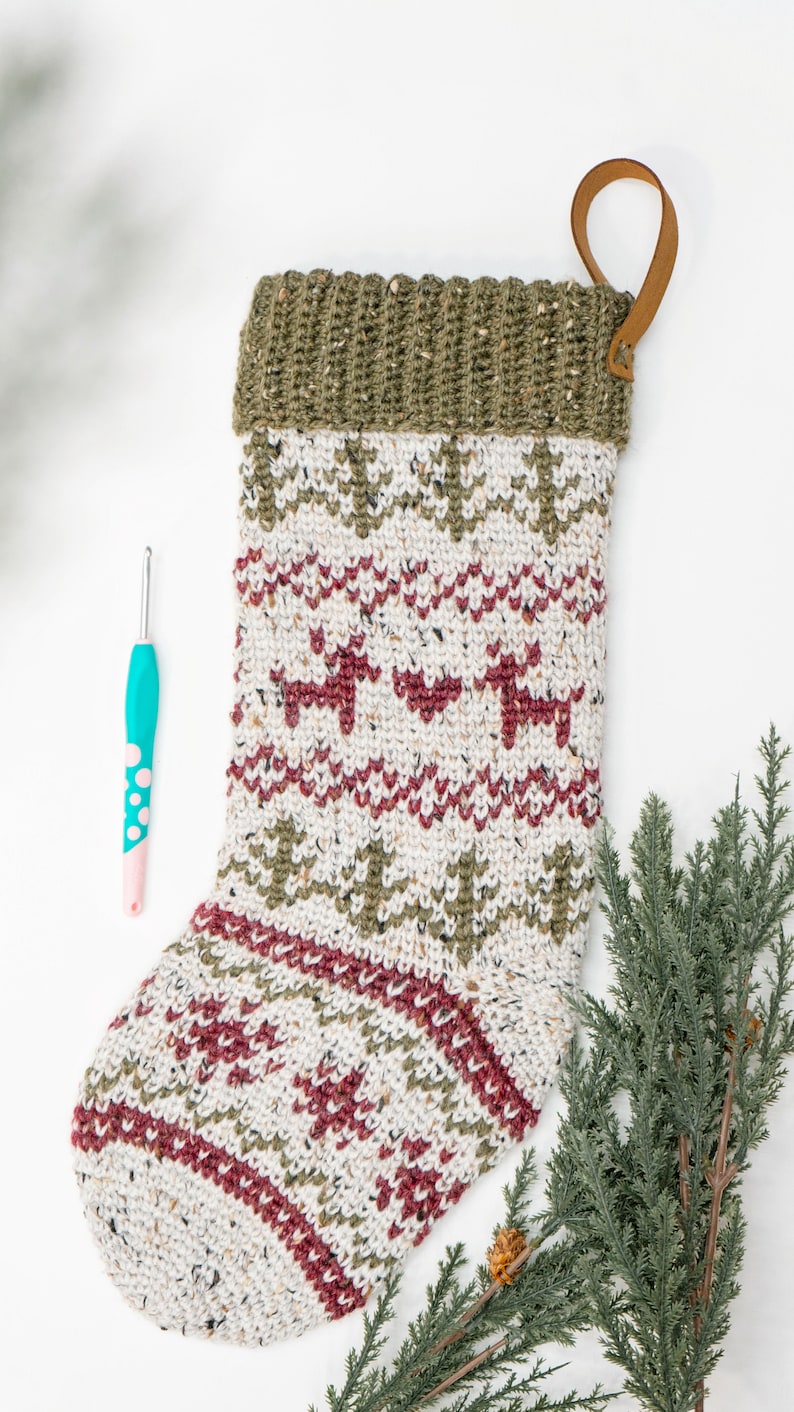 Op de afbeelding: Een gebreide kerstkous met een cr&egrave;mekleurige basis van gespikkeld garen, versierd met horizontale banden van donkerrode rendieren en olijfgroene dennenbomen, afgewerkt met een geribbelde olijfgroene boord en een bruine leren ophanglus.