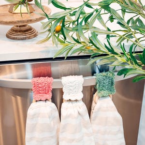 May include: Three kitchen towels with striped patterns hang from a stainless steel bar. Each towel has a crocheted topper in pink, white, and green. Olive branches with green leaves and small yellow fruits are in the background.