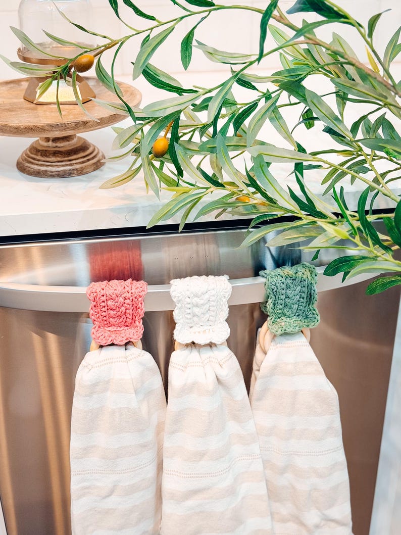 May include: Three kitchen towels with striped patterns hang from a stainless steel bar. Each towel has a crocheted topper in pink, white, and green. Olive branches with green leaves and small yellow fruits are in the background.