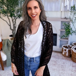 A woman with long gray hair, wearing a white top and blue jeans, holds up a large black crocheted shawl indoors—a perfect handmade gift—surrounded by plants and baskets in the background.