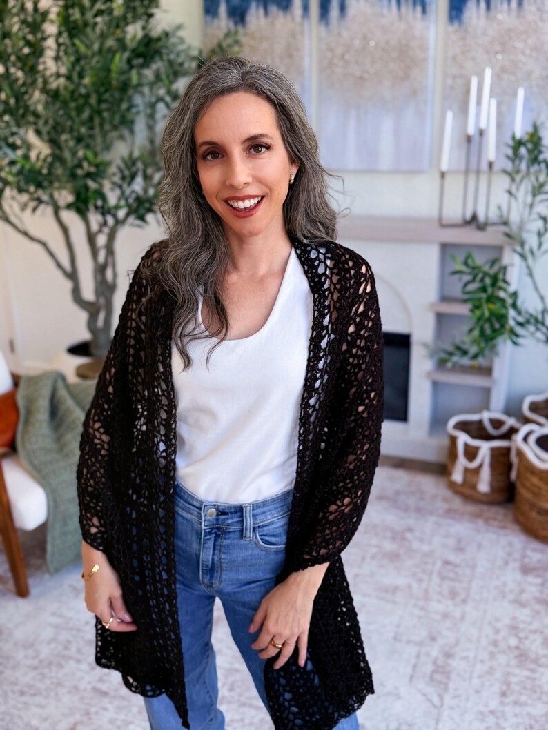A woman with long gray hair, wearing a white top and blue jeans, holds up a large black crocheted shawl indoors—a perfect handmade gift—surrounded by plants and baskets in the background.
