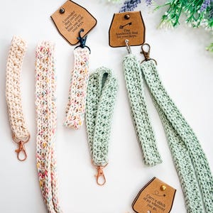 Puede incluir: Colección de llaveros de ganchillo hechos a mano en varios colores, incluyendo crema, arcoíris pastel y verde menta. Cada llavero tiene un cierre de mosquetón y una pequeña etiqueta con texto inspirador. Los llaveros se exhiben sobre una superficie blanca con detalles florales.