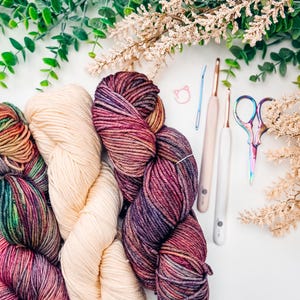 May include: Overhead view of colourful yarn skeins, crochet hooks, and scissors. The yarn is a mix of cream, purple, and multicoloured hues. The crochet hooks have white handles. The scissors have a rainbow finish. Greenery and white flowers are in the background.