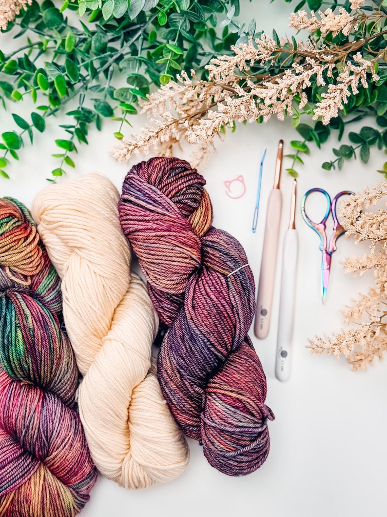 May include: Overhead view of colourful yarn skeins, crochet hooks, and scissors. The yarn is a mix of cream, purple, and multicoloured hues. The crochet hooks have white handles. The scissors have a rainbow finish. Greenery and white flowers are in the background.