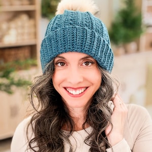 Teal crochet beanie with ribbed folded brim and cream pom pom worn by a smiling woman