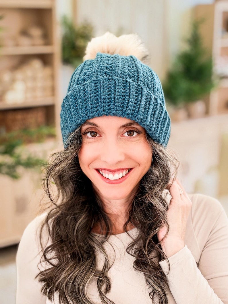 Teal crochet beanie with ribbed folded brim and cream pom pom worn by a smiling woman