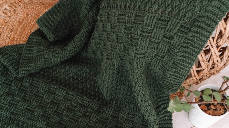 Puede incluir: Una manta tejida de color verde bosque oscuro que presenta un patr&oacute;n de textura de tejido de cesta. La tela muestra una construcci&oacute;n de puntadas densa y uniforme con un borde acanalado, exhibiendo un color profundo y s&oacute;lido en toda la pieza.