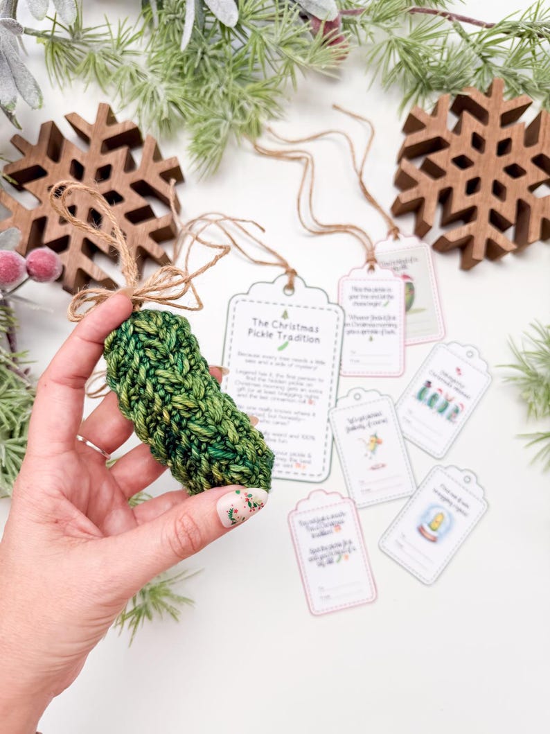 May include: A crocheted green pickle ornament with a jute twine loop, accompanied by several rectangular paper gift tags featuring printed text and illustrations related to the Christmas pickle tradition, including a larger instruction card.