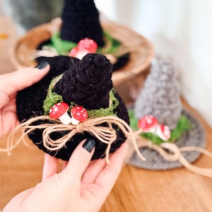 May include: A black crocheted witch hat featuring a green moss-like trim, two small red and white mushrooms, and a tan twine bow tied at the base. A second grey crocheted hat with similar decorations is visible in the background.