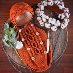 May include: A crocheted baby outfit in a burnt orange color with a matching crocheted hat. The outfit has a cable knit pattern and buttons down the front. The outfit is displayed on a wooden tray with a cotton wreath and a crochet hook.