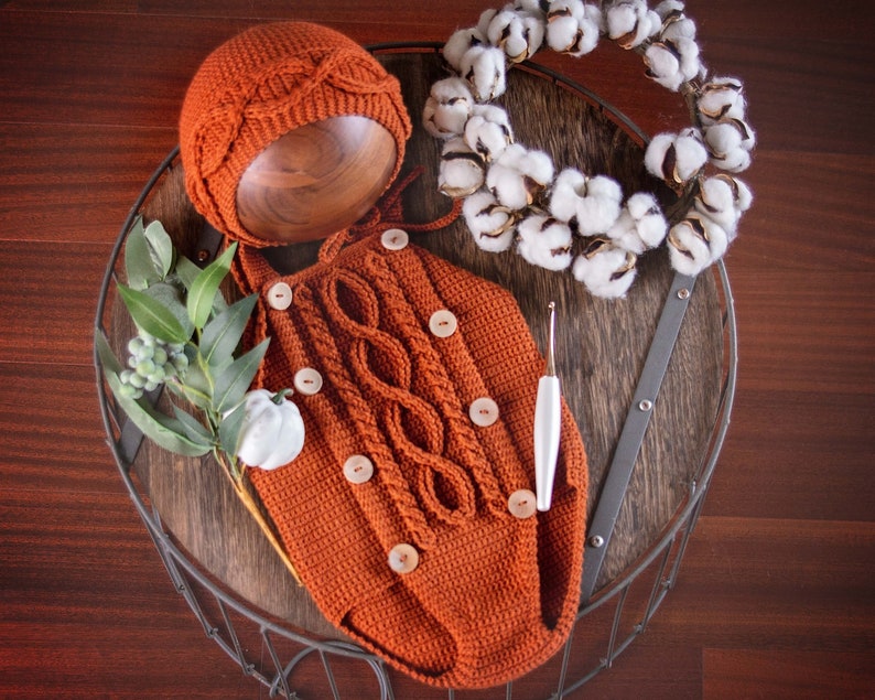 May include: A crocheted baby outfit in a burnt orange color with a matching crocheted hat. The outfit has a cable knit pattern and buttons down the front. The outfit is displayed on a wooden tray with a cotton wreath and a crochet hook.
