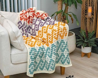 May include: A multi-colored crocheted throw blanket with a geometric pattern in shades of red, purple, yellow, and teal. The blanket is draped over a white armchair in a living room setting.