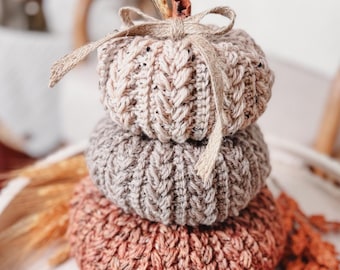 Op de afbeelding: Een stapel van drie gehaakte pompoenen in verschillende tinten crème, taupe en oranje. Elke pompoen heeft een gestructureerd kabelbreipatroon en is aan de bovenkant voorzien van een kleine bruine steel en een gestrikte jute touwtje.