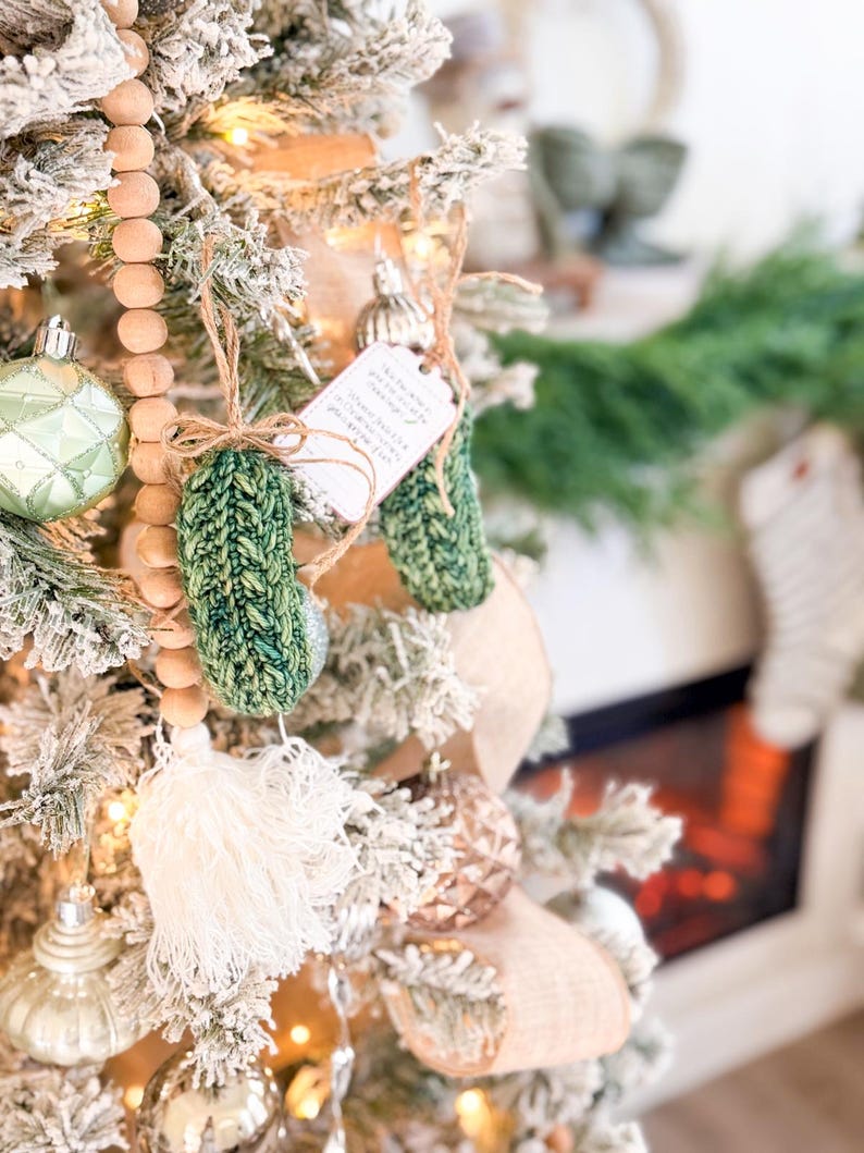 May include: A green knitted pickle ornament with a textured surface, hanging from a brown twine loop. A small white rectangular tag with printed text is attached to the twine. The ornament features a braided cable knit pattern in shades of green.