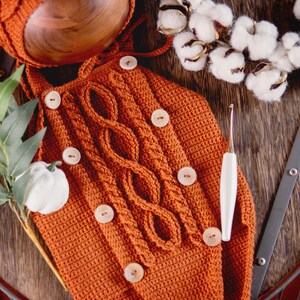 May include: A crocheted orange baby outfit with brown buttons and a white crochet hook. The outfit is laid out on a wooden surface with a sprig of greenery and a cotton boll wreath.