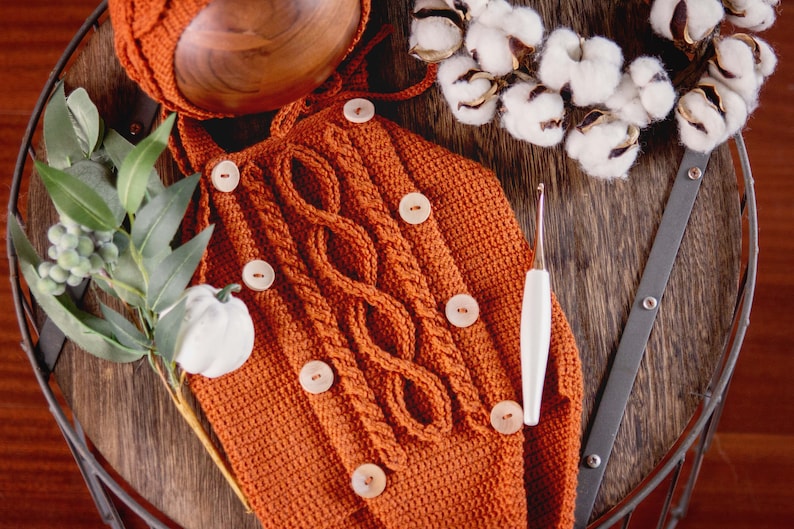 May include: A crocheted orange baby outfit with brown buttons and a white crochet hook. The outfit is laid out on a wooden surface with a sprig of greenery and a cotton boll wreath.