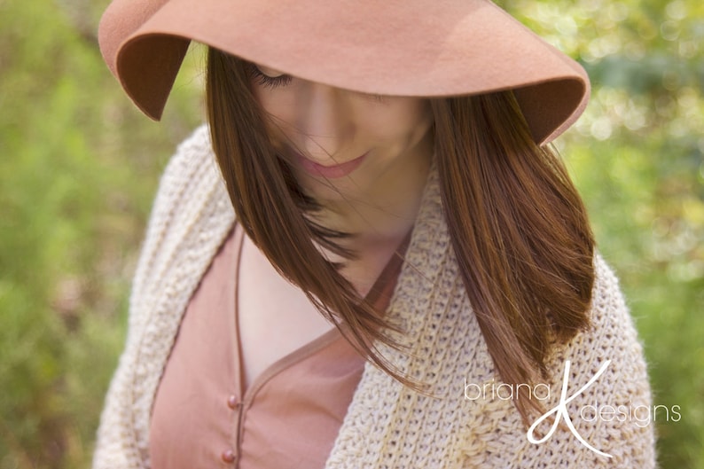 A woman wearing a cream crochet shawl wrap and hat, in a field of greenery.