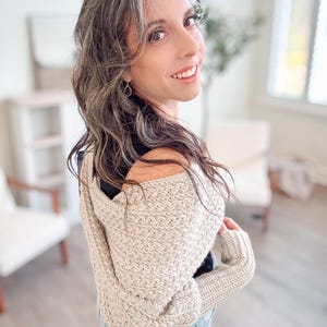 A woman with long hair and a light crochet sweater looks over her shoulder, smiling, in a bright room adorned with vintage clothes and a plant, exuding warmth and charm.