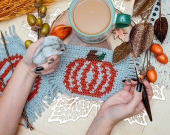 May include: A close-up of a crocheted pumpkin with orange and white yarn. The pumpkin is on a blue knitted blanket with fringe. A cup of coffee and a small hamster are also in the image.