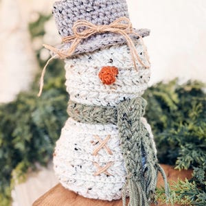 Speckled cream crochet snowman with gray hat, green fringed scarf and twine buttons on a wooden riser