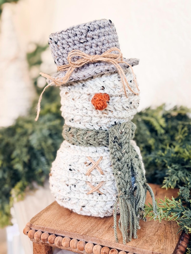 Speckled cream crochet snowman with gray hat, green fringed scarf and twine buttons on a wooden riser