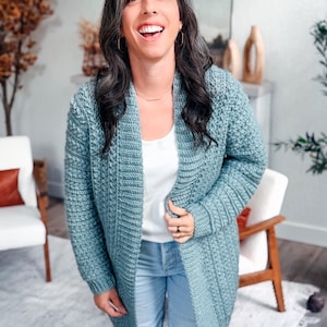 May include: A light blue crochet cardigan with a textured stitch pattern. The open-front cardigan is knee-length and worn over a white top and blue jeans. The cardigan has long sleeves and a relaxed fit, perfect for layering.