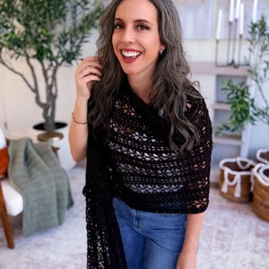 May include: A black crocheted shawl with an openwork pattern draped over the shoulders, paired with blue denim jeans and a white top. The shawl features a repeating geometric stitch design and hangs down on one side, showcasing the intricate texture.